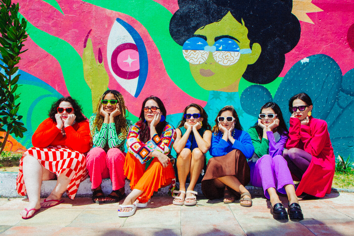 Grupo de mulheres sentadas em frente a um mural colorido, cada uma com cor e óculos diferentes